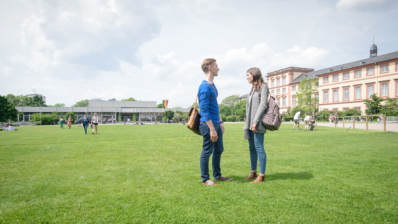 Eine Studentin und ein Student stehen auf der Mensawiese der Universität Mannheim und unterhalten sich.