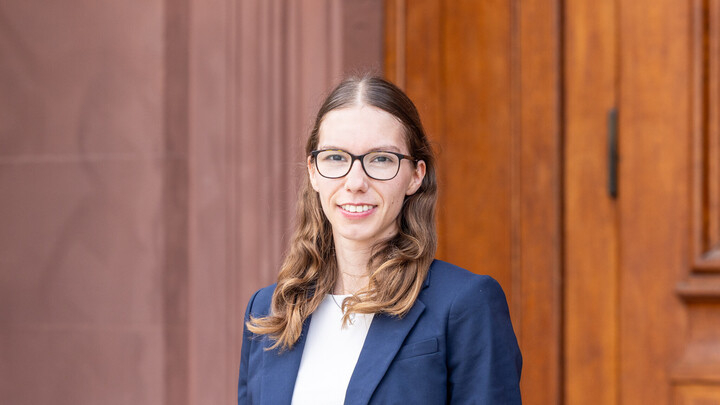 A portrait of Maren Fritz. She is wearing a dark blue blazer and standing in front of a door in the castle.