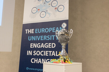 Ein silberner Pokal steht auf einem Regal. Hinter dem Regal steht ein Banner, auf dem folgendes zu lesen ist: "The european university engaged in societal changes".
