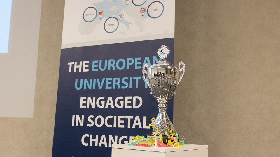 Ein silberner Pokal steht auf einem Regal. Hinter dem Regal steht ein Banner, auf dem folgendes zu lesen ist: "The european university engaged in societal changes".