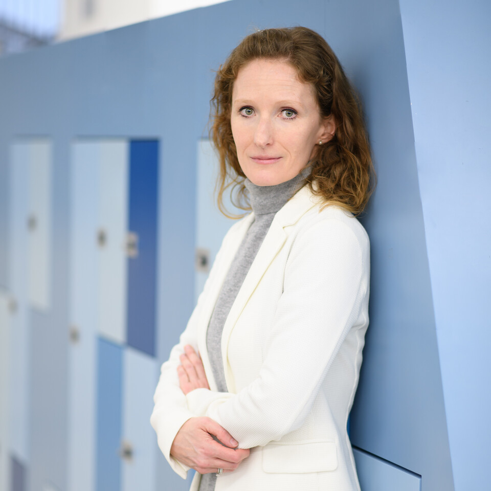 Porträt von Karin Hoisl. Sie hat braune, halblange Haare, trägt ein weißes Jacket und schaut in die Kamera. Sie lehnt an einer Wand mit blauen Schließfächern.