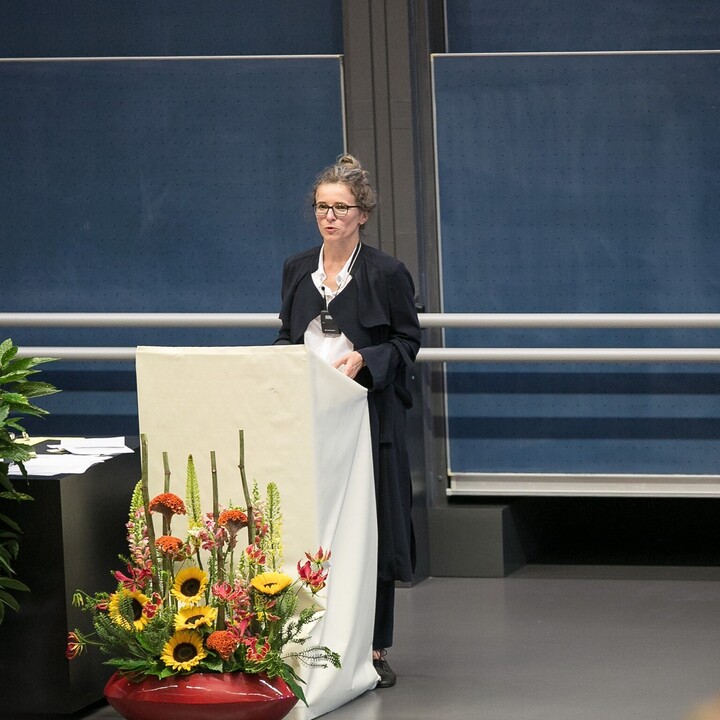 Eine Person trägt eine weiße Bluse sowie einen schwarzen Mantel und steht hinter einem Rednerpult in einem Hörsaal. Die Person heißt Melanie Meder. 