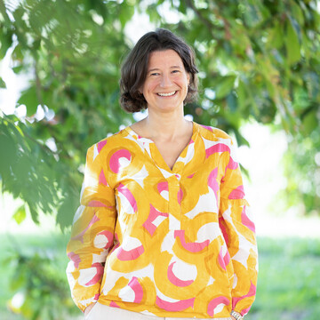 Dr. Katrin Schoppa-Bauer. Sie steht locker da und lächelt. Sie trägt eine rosa und gelb gemusterte Bluse.