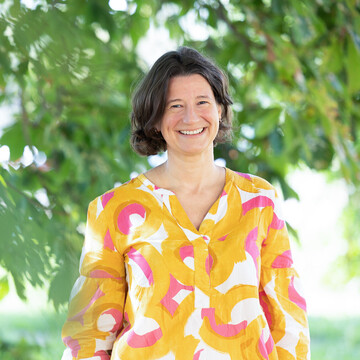 Dr. Katrin Schoppa-Bauer. Sie steht locker da und lächelt. Sie trägt eine rosa und gelb gemusterte Bluse.