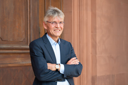 Professor Spengel trägt einen Anzug und hat die Arme ineinander verschränkt. Er steht vor einer Holztür.
