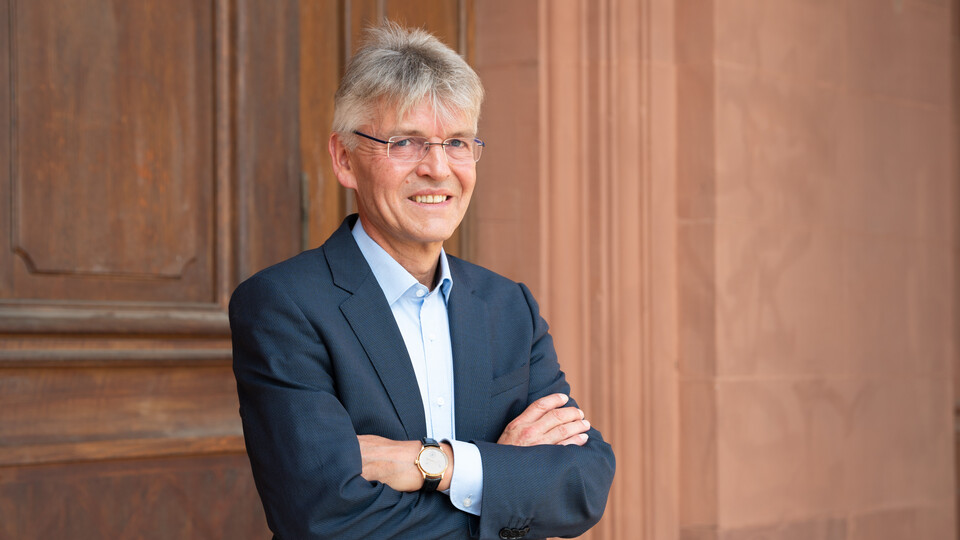 Professor Spengel trägt einen Anzug und hat die Arme ineinander verschränkt. Er steht vor einer Holztür.