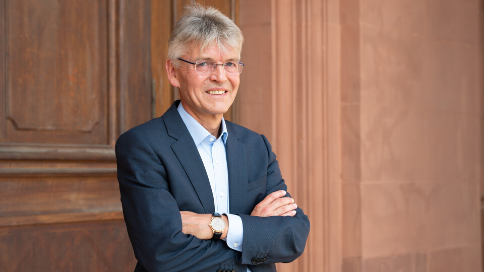 Professor Spengel trägt einen Anzug und hat die Arme ineinander verschränkt. Er steht vor einer Holztür.