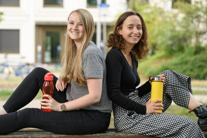 Zwei Studentinnen lehnen Rücken an Rücken draußen auf einer Bank. Sie haben beide eine Trinkflasche in der Hand.