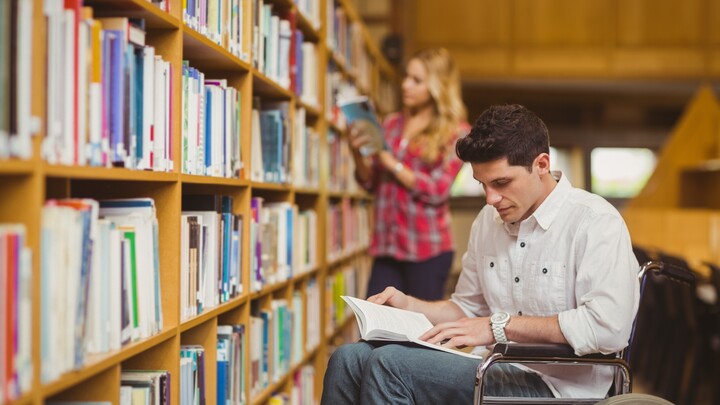 Ein Studierender im Rollstuhl mit einem Buch auf dem Schoß vor einem Bücherregal. Link: Beratung für behinderte und chronisch kranke Studierende