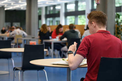 Ein Student in rotem Tshirt sitzt in der Mensa an einem Tisch und isst zu Mittag.