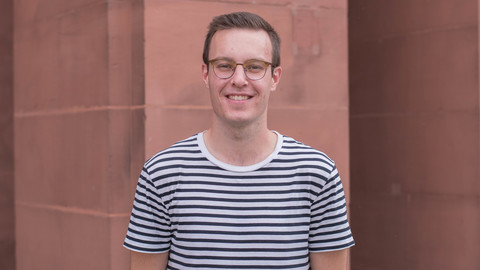 Nico Kasparetz. He is wearing a black-white stripped T-shirt and is standing in front of a brown stone wall.