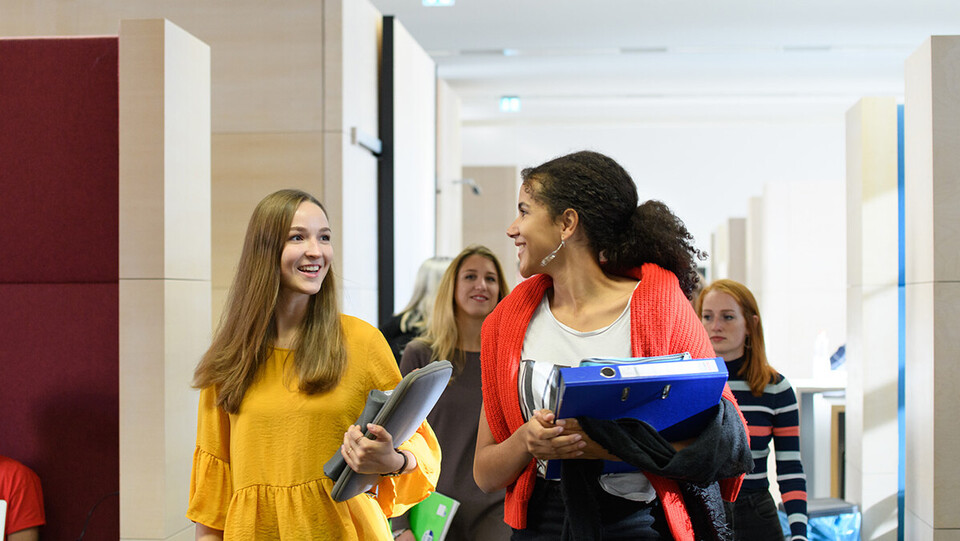 Eine Gruppe weiblicher Studierender mit Lernsachen in den Händen läuft durch die Bibliothek und unterhält sich