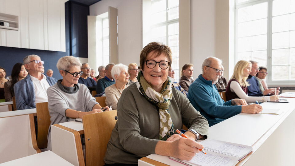 Mehrere Menschen in höherem Alter sitzen in einem Hörsaal. Eine Person lächelt in die Kamera.