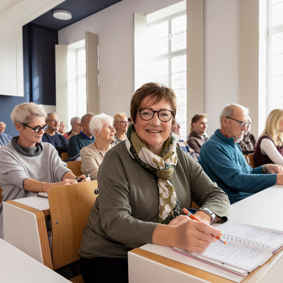 Mehrere Menschen in höherem Alter sitzen in einem Hörsaal. Eine Person lächelt in die Kamera.
