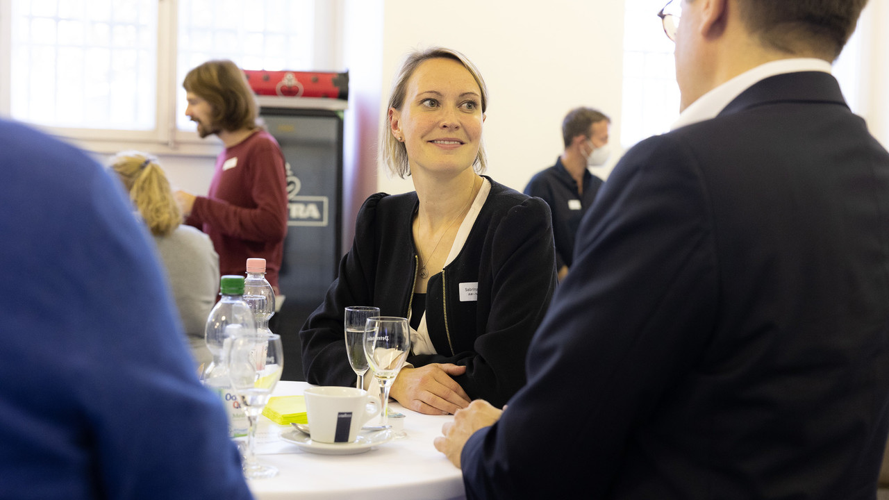 Eine Frau unterhält sich angeregt an einem Bistro-Tisch, auf dem Sektgläser, Kaffeetassen und Wasserflaschen stehen.