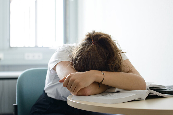 Eine Studierende liegt ermattet mit ihrem Kopf und verschränkten Armen auf ihren Büchern.