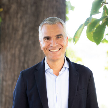  Prof. Dr. Thomas Fetzer. Breit lächelnd steht er vor einem Baum, dabei trägt er ein dunkles Jackett.