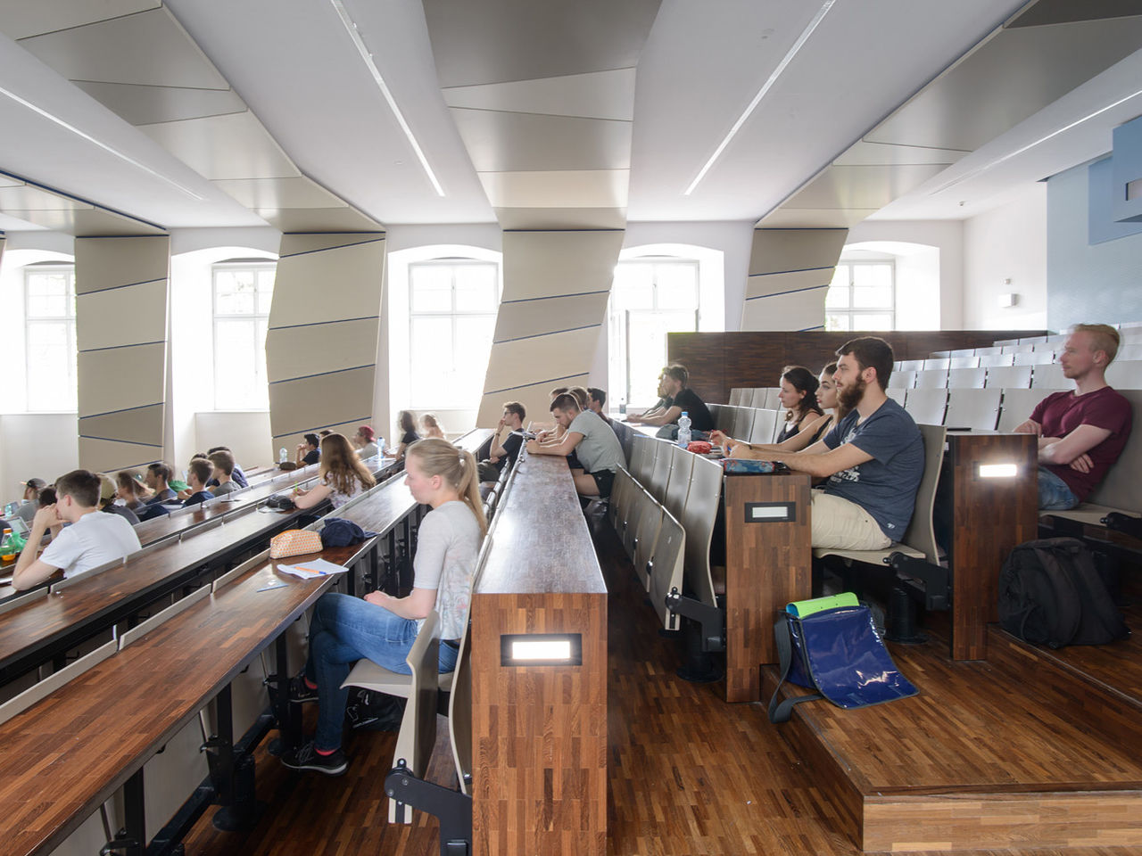 Studierende in einem hölzernen, lichtdurchfluteten Hörsaal mit vielen Fenstern