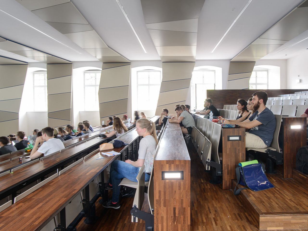 Studierende in einem hölzernen, lichtdurchfluteten Hörsaal mit vielen Fenstern