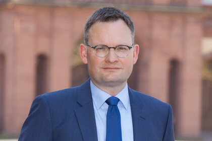 Eine Person trägt ein hellblaues Hemd, eine blaue Krawatte sowie einen dunkelblauen Blazer und steht vor dem Schloss der Universität Mannheim. Die Person heißt Philipp Gassert.