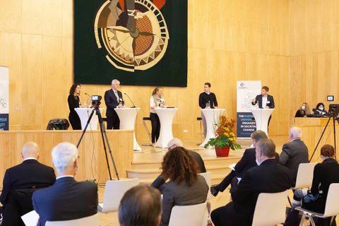 In einem Vorlesungssaal stehen fünf Personen in schicker Kleidung an jeweils einem Stehtisch. Sie stehen vor einem Publikum, das auf Stühlen sitzt. 