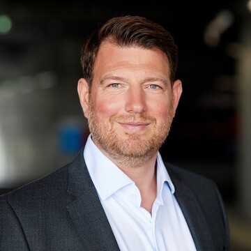 Philipp Laux hat braune Haare und einen grau-braunen Dreitagebart. Er trägt ein graues Jacket und ein weißes Hemd.