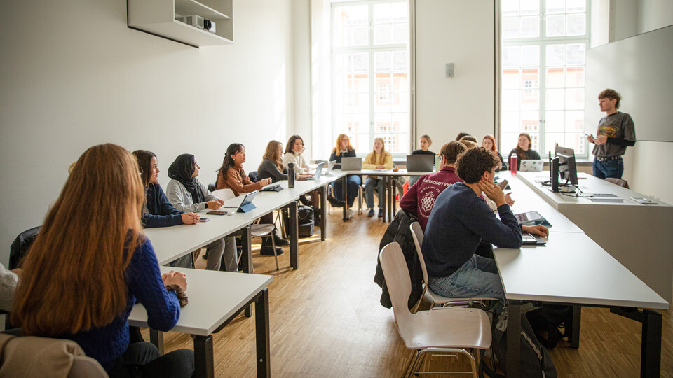 Studierende sitzen in einem Seminarraum an Tischen und schauen nach vorne zum Dozierenden.