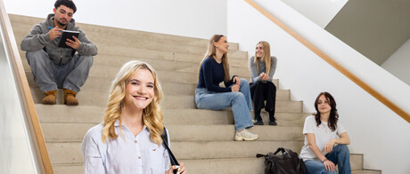 Eine Studentin mit einer hellen Bluse, einer dunklen Tasche und blonden Haaren steht im B6 Gebäude vor einer Treppe. Auf der Treppe sitzen und stehen im Hintergrund 5 Studierende.