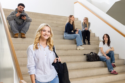 Eine Studentin mit einer hellen Bluse, einer dunklen Tasche und blonden Haaren steht im B6 Gebäude vor einer Treppe. Auf der Treppe sitzen und stehen im Hintergrund 5 Studierende.