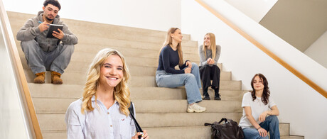 Eine Studentin mit einer hellen Bluse, einer dunklen Tasche und blonden Haaren steht im B6 Gebäude vor einer Treppe. Auf der Treppe sitzen und stehen im Hintergrund 5 Studierende.