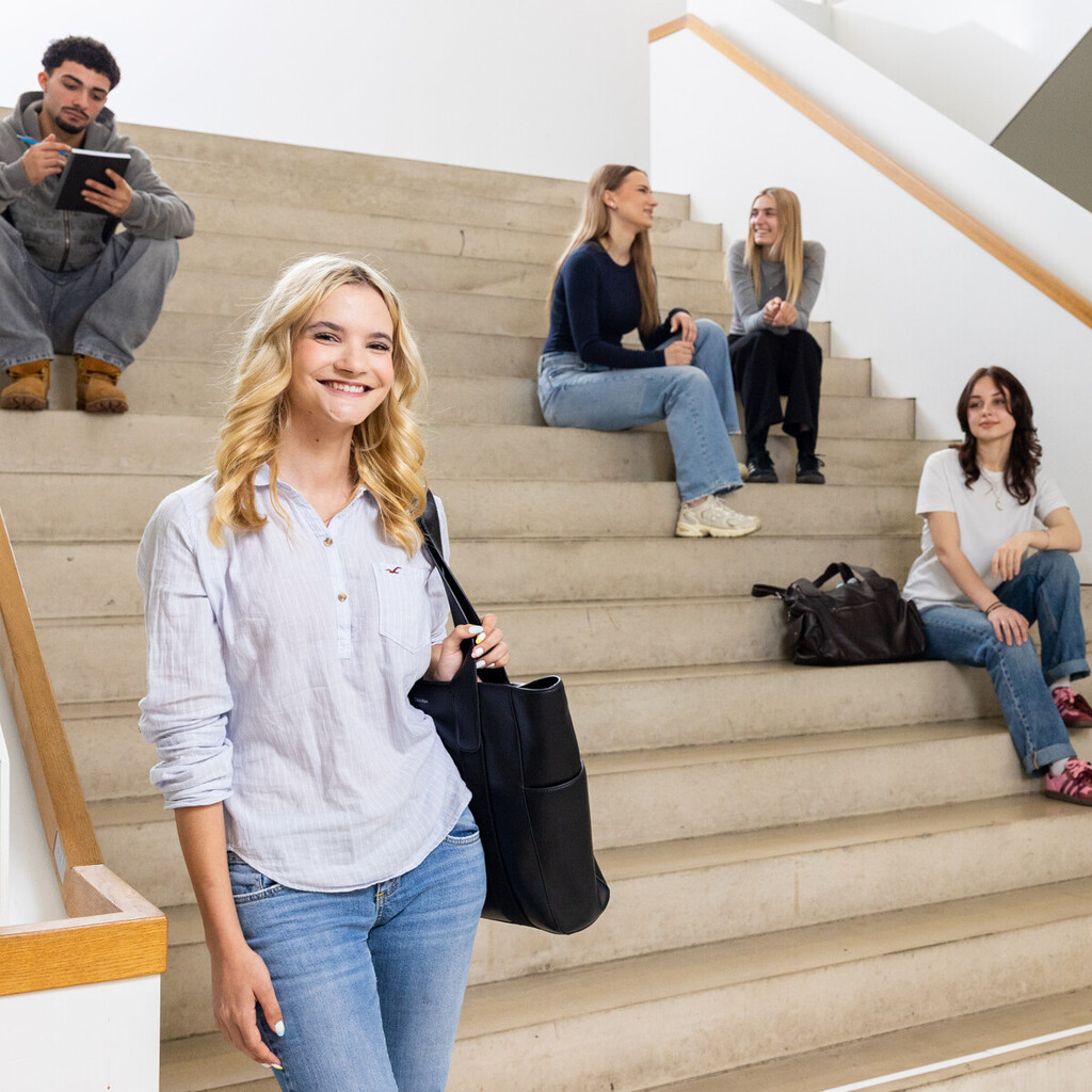 Eine Studentin mit einer hellen Bluse, einer dunklen Tasche und blonden Haaren steht im B6 Gebäude vor einer Treppe. Auf der Treppe sitzen und stehen im Hintergrund 5 Studierende.