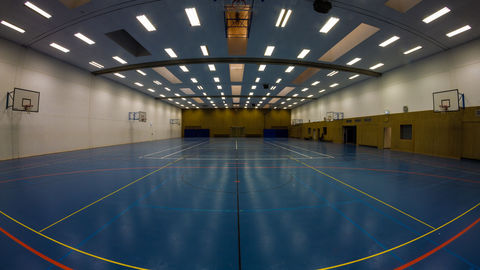 Hellbestrahlte Unihalle mit blauem Gummiboden, der durch bunte Linien in verschiedene Felder eingeteilt ist. Die Decke ist ebenfalls blau und mit Neonleuchten versehen. An den weißen Seitenwänden hängen zwei Basketballkörbe.