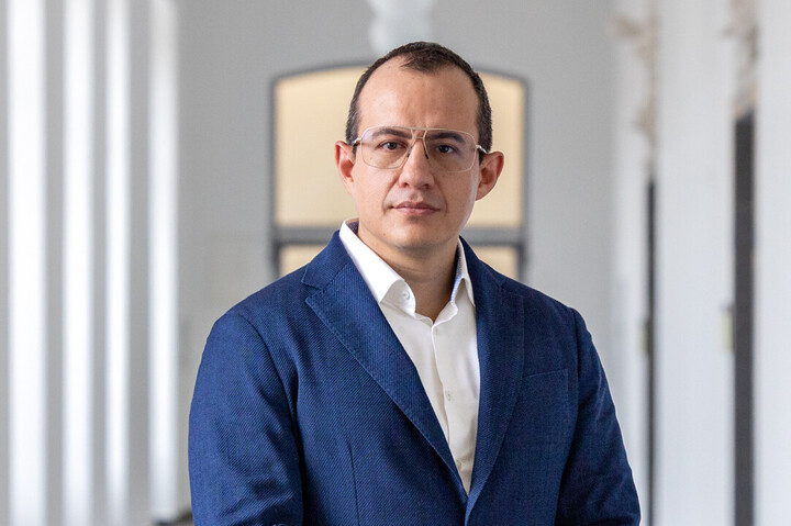 Alumnus José Márquez hat kurze, dunkle Haare und trägt eine Brille. Er hat ein weißes Hemd an mit einem blauen Sakko darüber und steht in einem langen Flur.