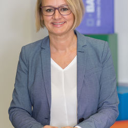 Frau mit blonden Haaren und einem Bobschnitt. Sie trägt eine weiße Bluse, einen mit sehr kleinen weißen Punkten versehenen blauen Blazer, eine Silberuhr sowie eine Brille. Sie lächelt, steht an einem Glastisch und scheint seriös zu sein.