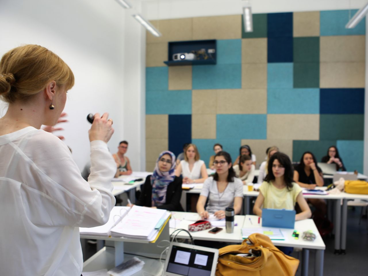 Eine Dozentin mit weißer Bluse und blonden zusammengebundenen Haaren spricht vor Studierenden im Seminarraum.