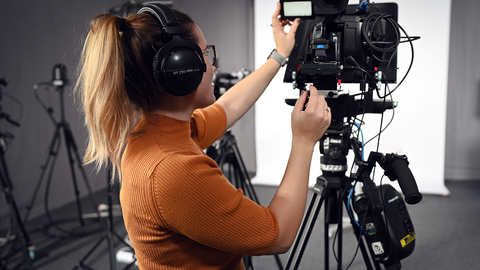 Eine Person trägt ein orange-farbenes T-Shirt sowie Kopfhörer und steht in einem Videostudio. Die Person steht hinter einer Kamera und blickt auf diese.