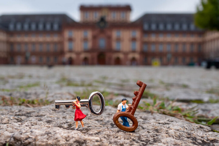 Zwei Miniaturfiguren, eine Frau im roten Kleid und ein Mann in blauer Latzhose, stehen auf Kopfsteinpflaster und tragen übergroße Schlüssel; im Hintergrund ist unscharf das Mannheimer Schloss zu erkennen.
