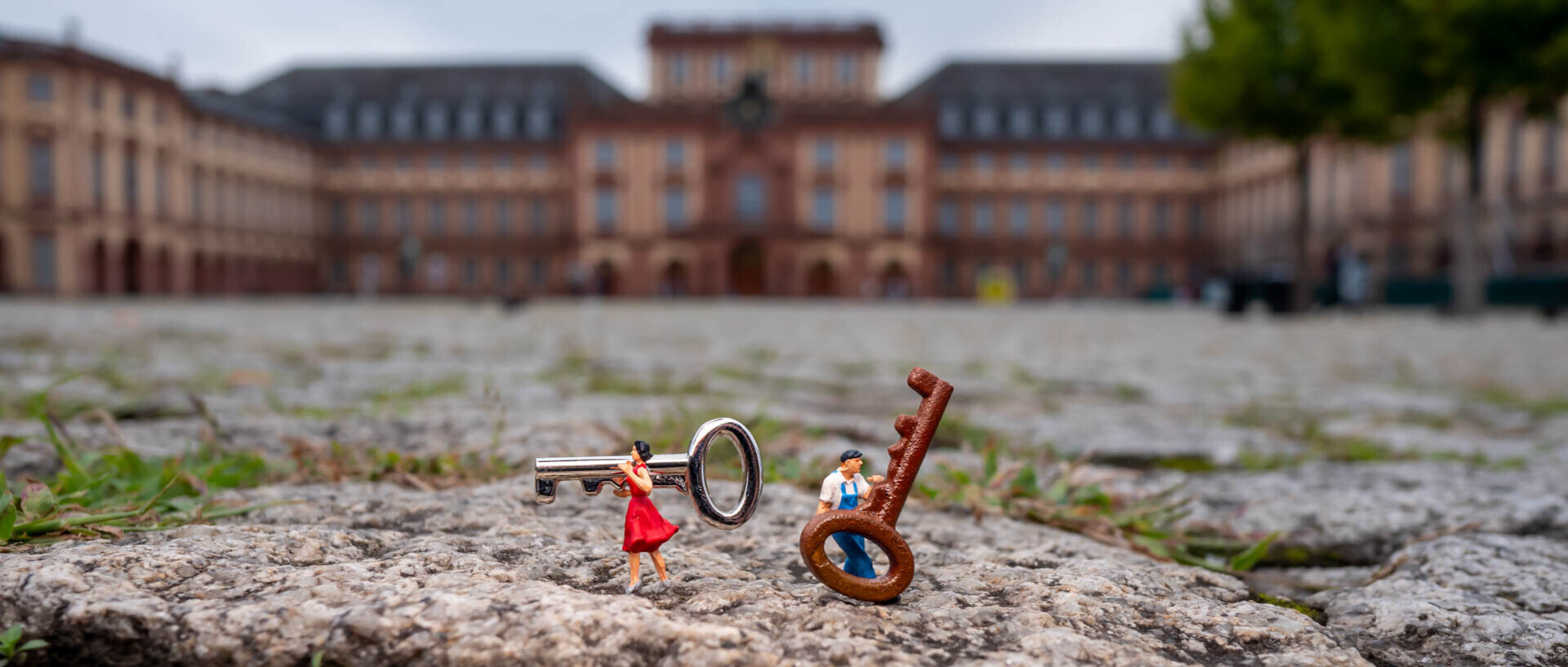 Zwei Miniaturfiguren, eine Frau im roten Kleid und ein Mann in blauer Latzhose, stehen auf Kopfsteinpflaster und tragen übergroße Schlüssel; im Hintergrund ist unscharf das Mannheimer Schloss zu erkennen.