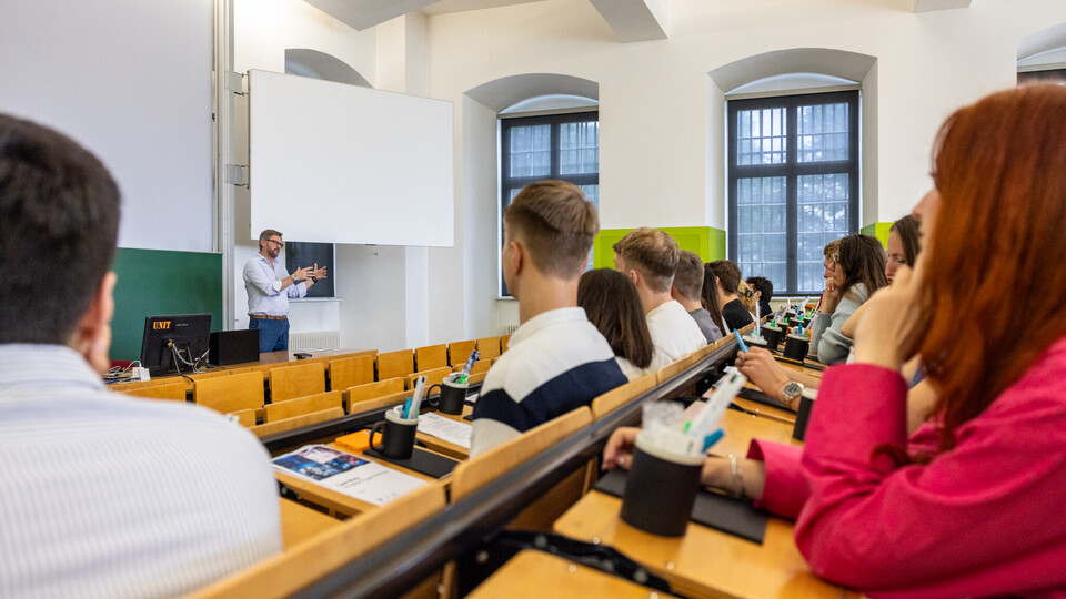 In einem Hörsaal sitzen Studierende. Ein Mann steht vorne und redet.