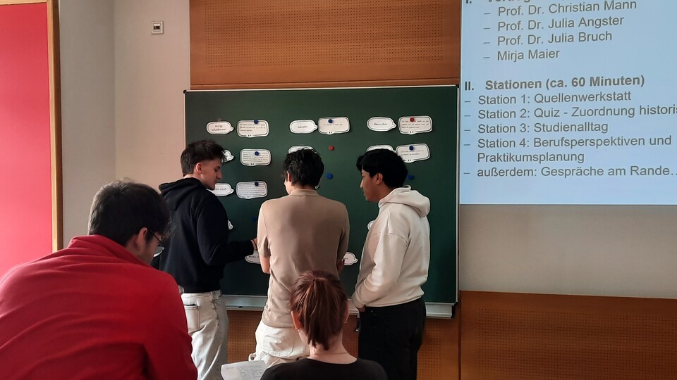 Drei Studierende stehen vor einer Tafel mit mehreren Zetteln darauf und unterhalten sich. 