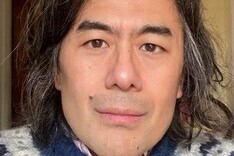 Charles Yang hat mittellange Haare mit grauen Strähnen, einen Leberfleck über dem rechten Mundwinkel und einen ernsten Blick. Er trägt ein Hemd unter einem Wollpulli
