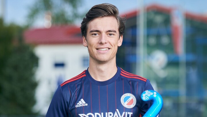 Raphael Hartkopf steht auf einem Hockeyfeld. Er trägt rote Shorts und ein blaues Trikot. Er lächelt in die Kamera. In der linken Hand hält er einen Hockeyschläger.