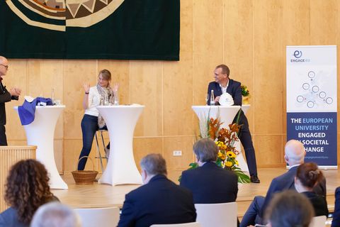 Drei Personen in schicker Kleidung sitzen hinter jeweils einem Stehtisch und unterhalten sich. Im Vordergrund sitzt ein Publikum.