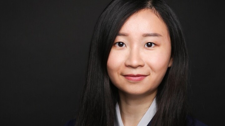 Mengnan Wu. She is wearing a black jacket over a white shirt and is standing in front of black background.