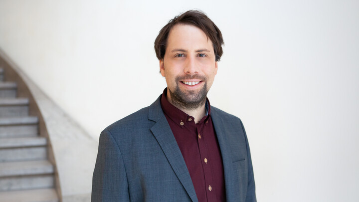 Eine lächelnde Person trägt ein weinrotes Hemd sowie einen grauen Blazer und steht vor einer weißen Wand. Die Person heißt Tobias Ebert.