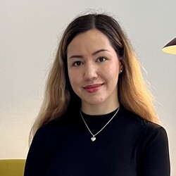 Tsvetina Tsonkova has light brown hair, which is worn loose. She is sitting on a green armchair and is wearing a black turtleneck sweater and jeans.