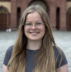 Vanessa Schütz hat lange blonde Haare und lacht in die Kamera. Sie trägt eine große Brille mit goldenem gestell und ein lila-graues Top.