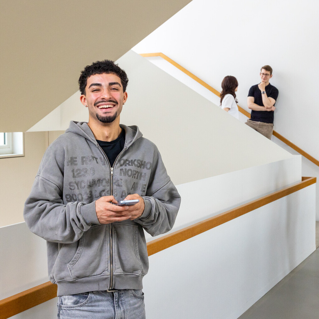 Ein Student steht im Treppenhaus des B6 Gebäudes. Er hat dunkle Haare, trägt einen grauen Hoodie und lacht. Er hat ein Handy in der Hand. 
