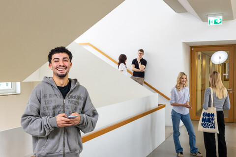 Ein Student steht im Treppenhaus des B6 Gebäudes. Er hat dunkle Haare, trägt einen grauen Hoodie und lacht. Er hat ein Handy in der Hand. 