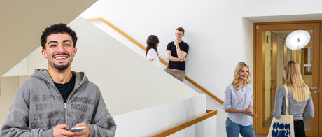 Ein Student steht im Treppenhaus des B6 Gebäudes. Er hat dunkle Haare, trägt einen grauen Hoodie und lacht. Er hat ein Handy in der Hand. 